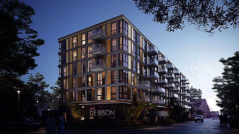 Impressie van modern appartementencomplex "De Bison" in een groene wijk bij schemering, met verlichte ramen en glazen balkons.